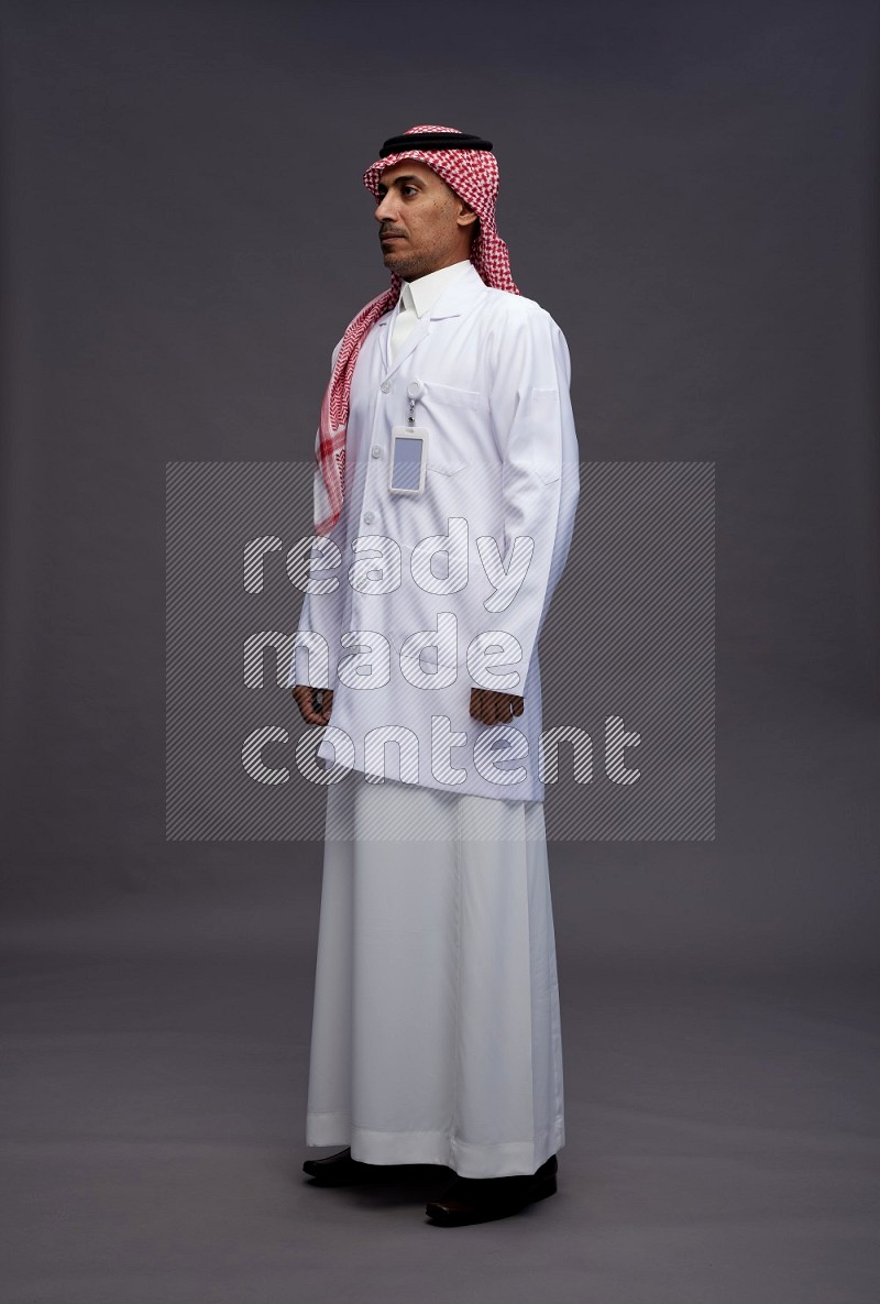 Saudi man wearing thob with lab coat and shomag with pocket employee badge standing interacting with the camera on gray background