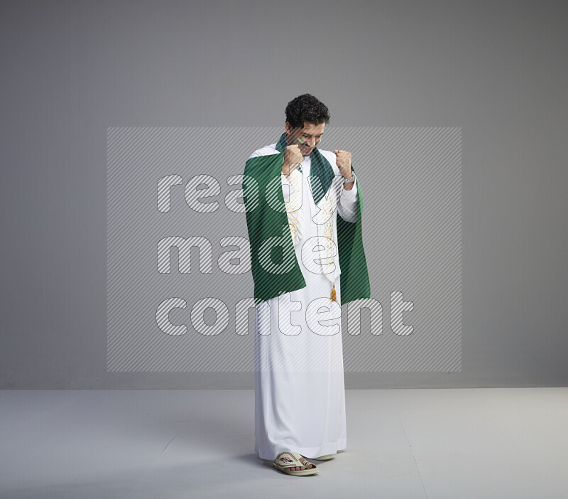 A Saudi man standing wearing thob with face painting wrapping Saudi flag scarf and holding big Saudi flag on gray background