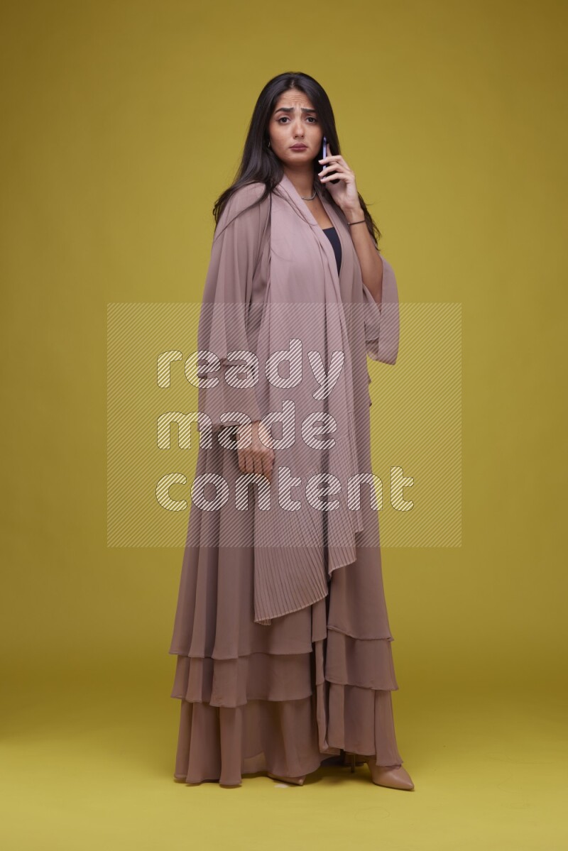 A woman Calling on a Yellow Background wearing Brown Abaya