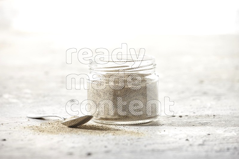 A glass jar and a metal spoon full of white pepper powder on textured white flooring