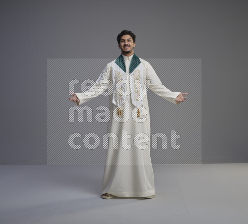 A Saudi man standing wearing thob and Saudi flag scarf on gray background