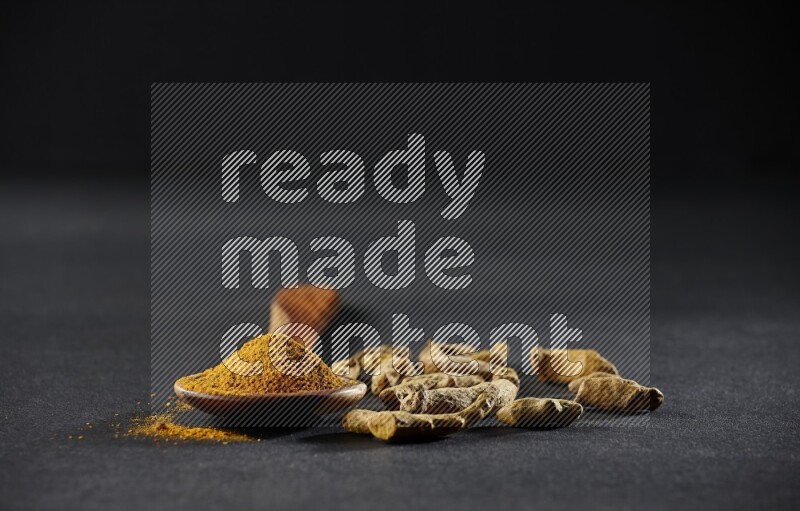 A wooden ladle full of turmeric powder with dried turmeric fingers on black flooring