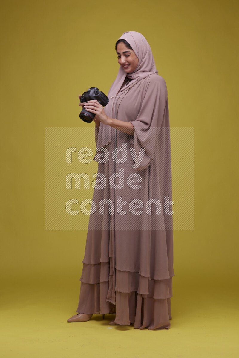 A woman Holding a Camera on a Yellow Background wearing Brown Abaya with Hijab