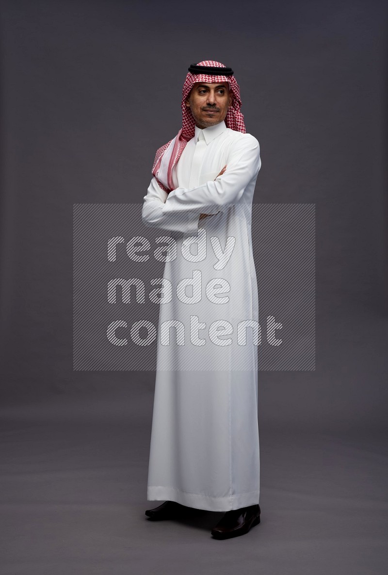 Saudi man wearing thob and shomag standing with crossed arms on gray background