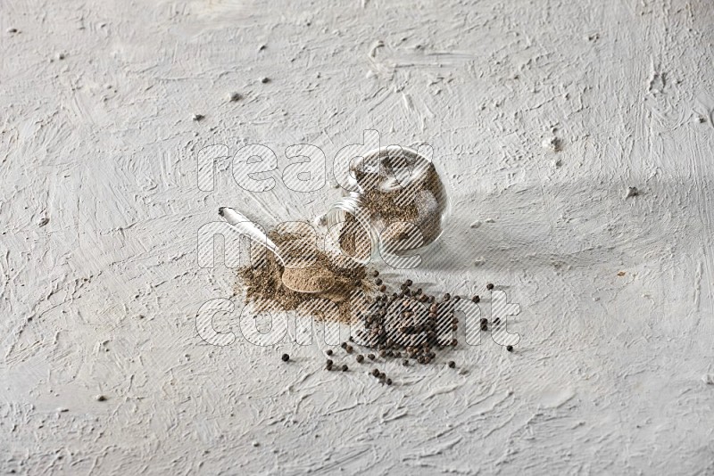 Flipped glass spice jar full of black pepper powder with a metal spoon and black pepper beads spread on a textured white flooring