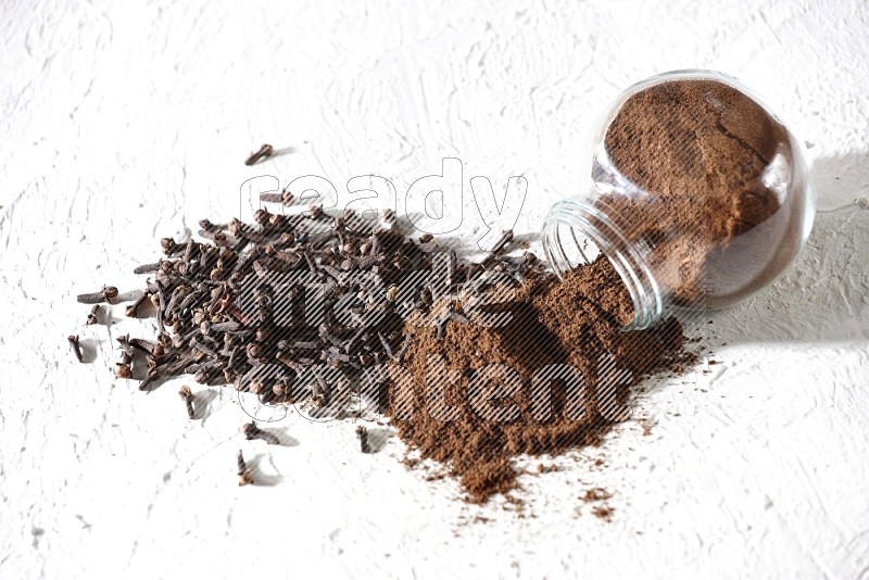 A glass spice jar full of cloves powder flipped and powder came out of it with cloves spread on textured white flooring