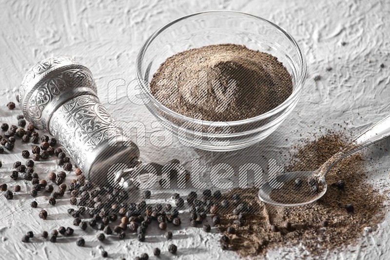 A glass bowl full of black pepper powder with black pepper beads, a turkish metal grinder and a metal spoon on textured white flooring