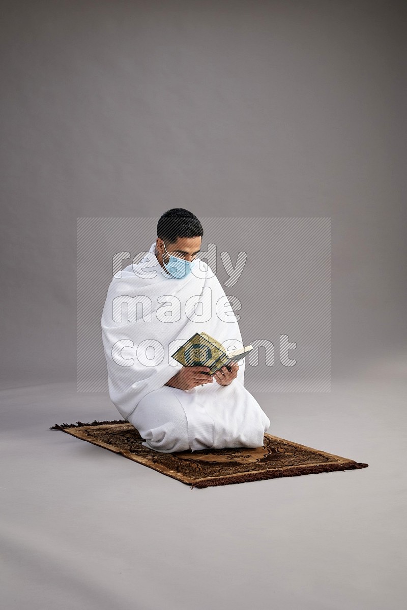 A man wearing Ehram with face mask sitting on floor reading quran on gray background