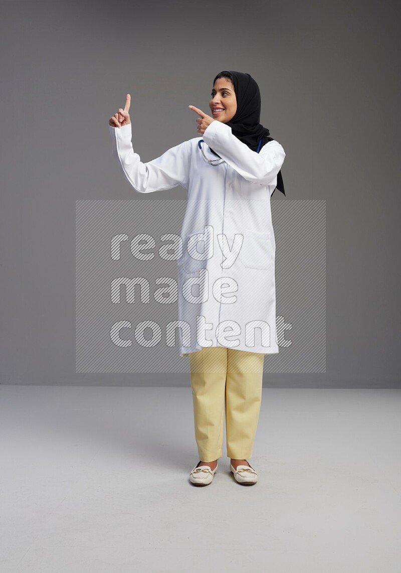Saudi woman wearing lab coat with stethoscope standing interacting with the camera on Gray background