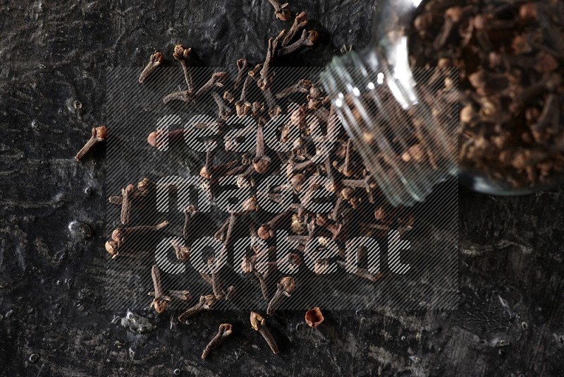 A flipped glass spice jar full of cloves and cloves came out of it on textured black flooring