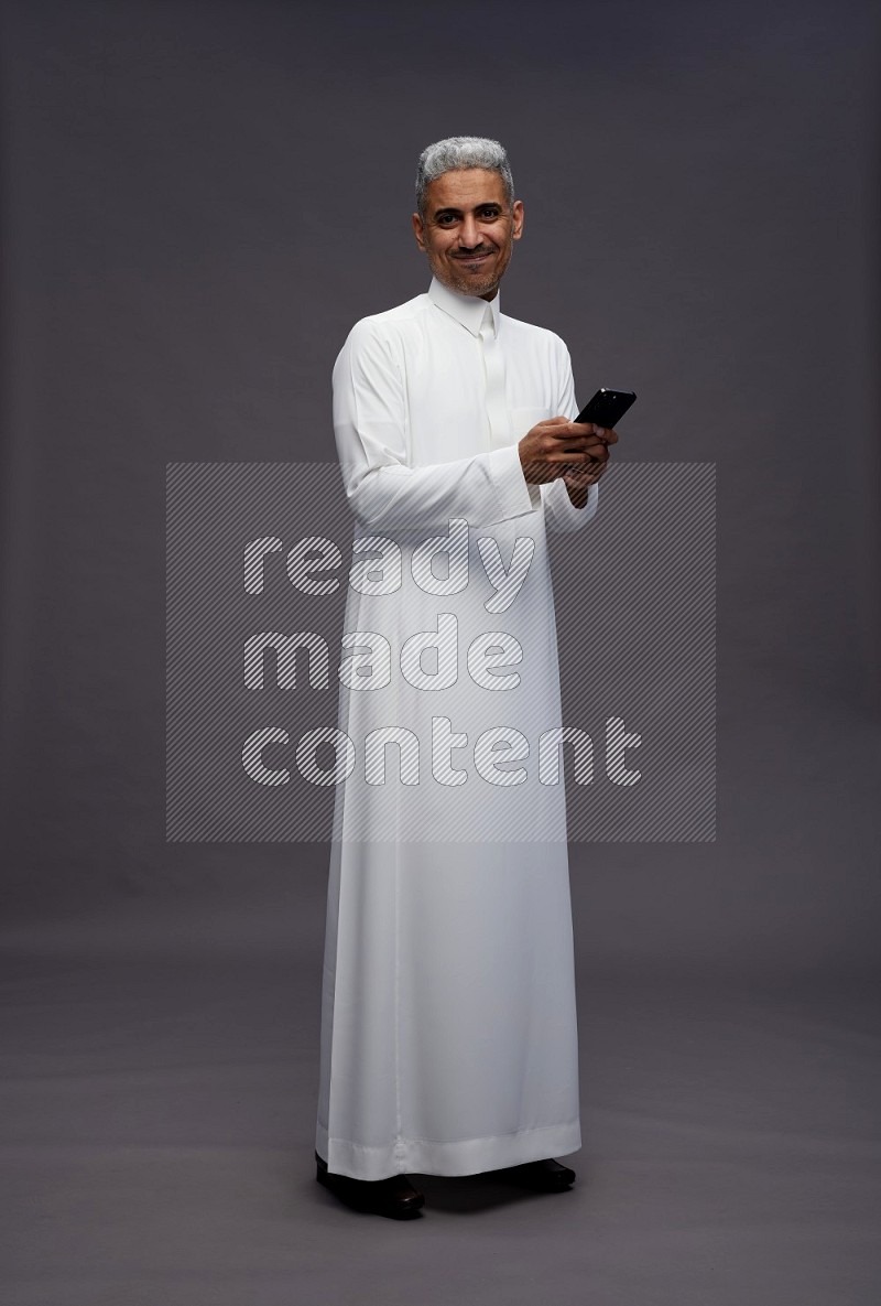 Saudi man wearing thob standing texting on phone on gray background