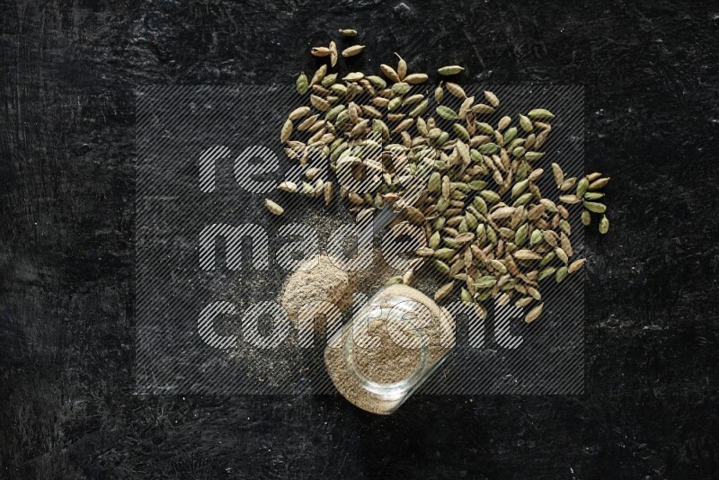 A glass spice jar and a metal spoon full of cardamom powder and cardamom seeds spreaded on textured black flooring
