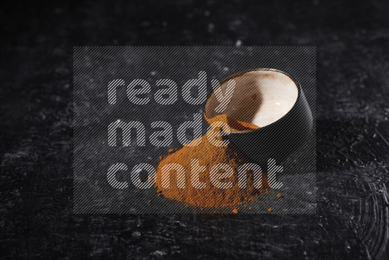 A black pottery bowl full of ground paprika powder with fallen powder from it on black background