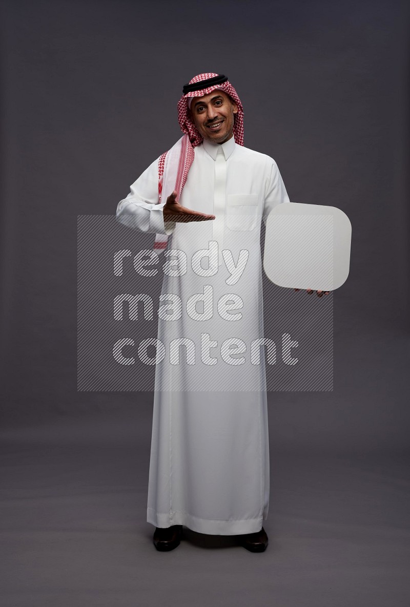 Saudi man wearing thob and shomag standing holding social media sign on gray background