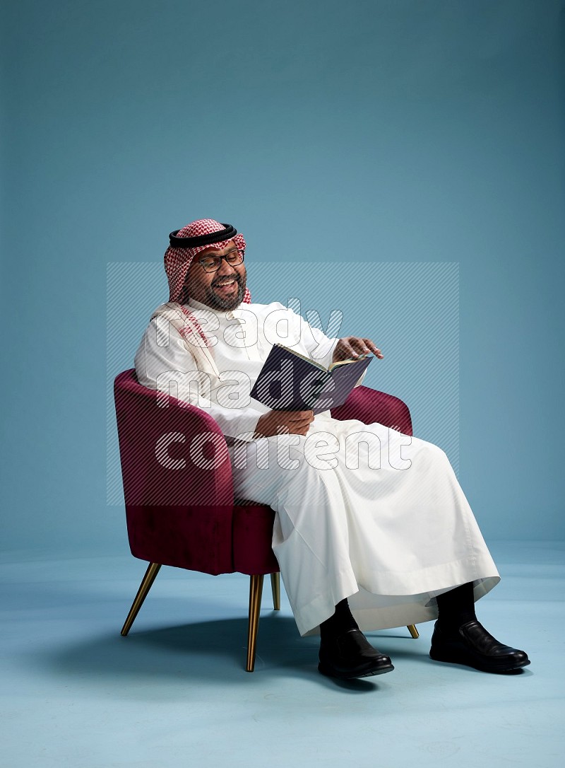 Saudi Man with shimag sitting on chair reading book on blue background