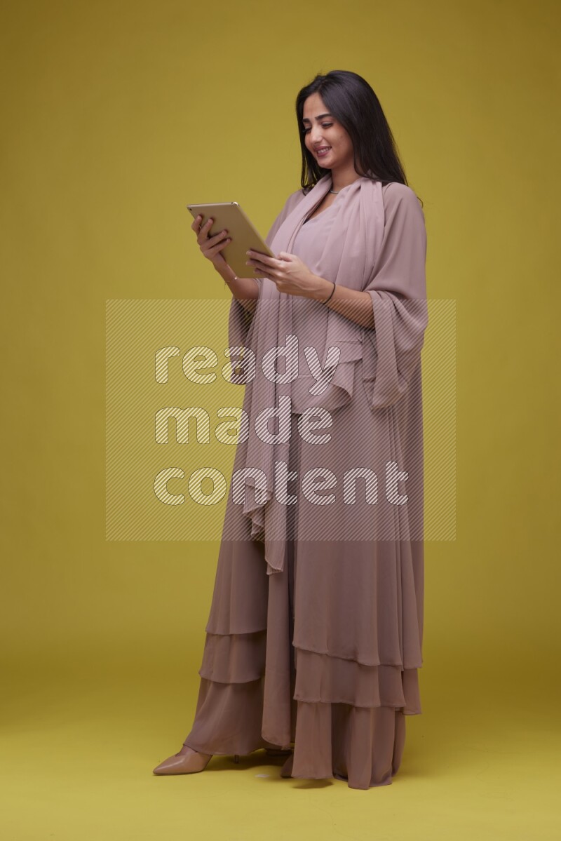 A Saudi woman Holding an iPad  on a Yellow Background wearing Brown Abaya