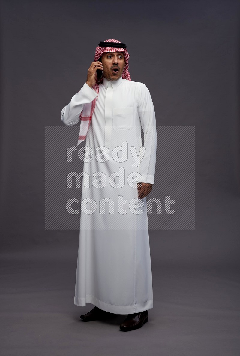 Saudi man wearing thob and shomag standing talking on phone on gray background