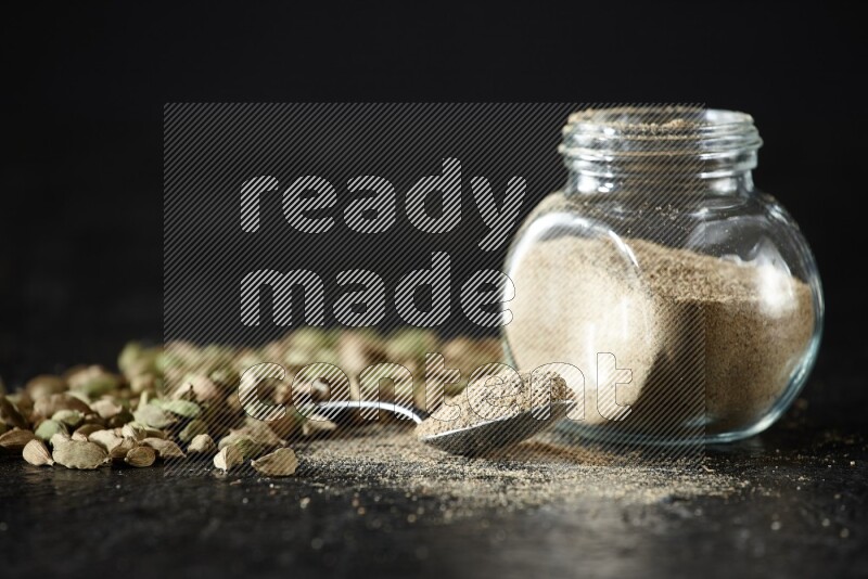 A glass spice jar and a metal spoon full of cardamom powder and cardamom seeds spreaded on textured black flooring