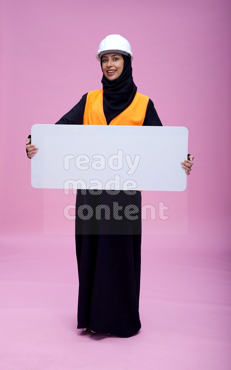 Saudi woman wearing Abaya with engineer vest and helmet standing holding board on pink background