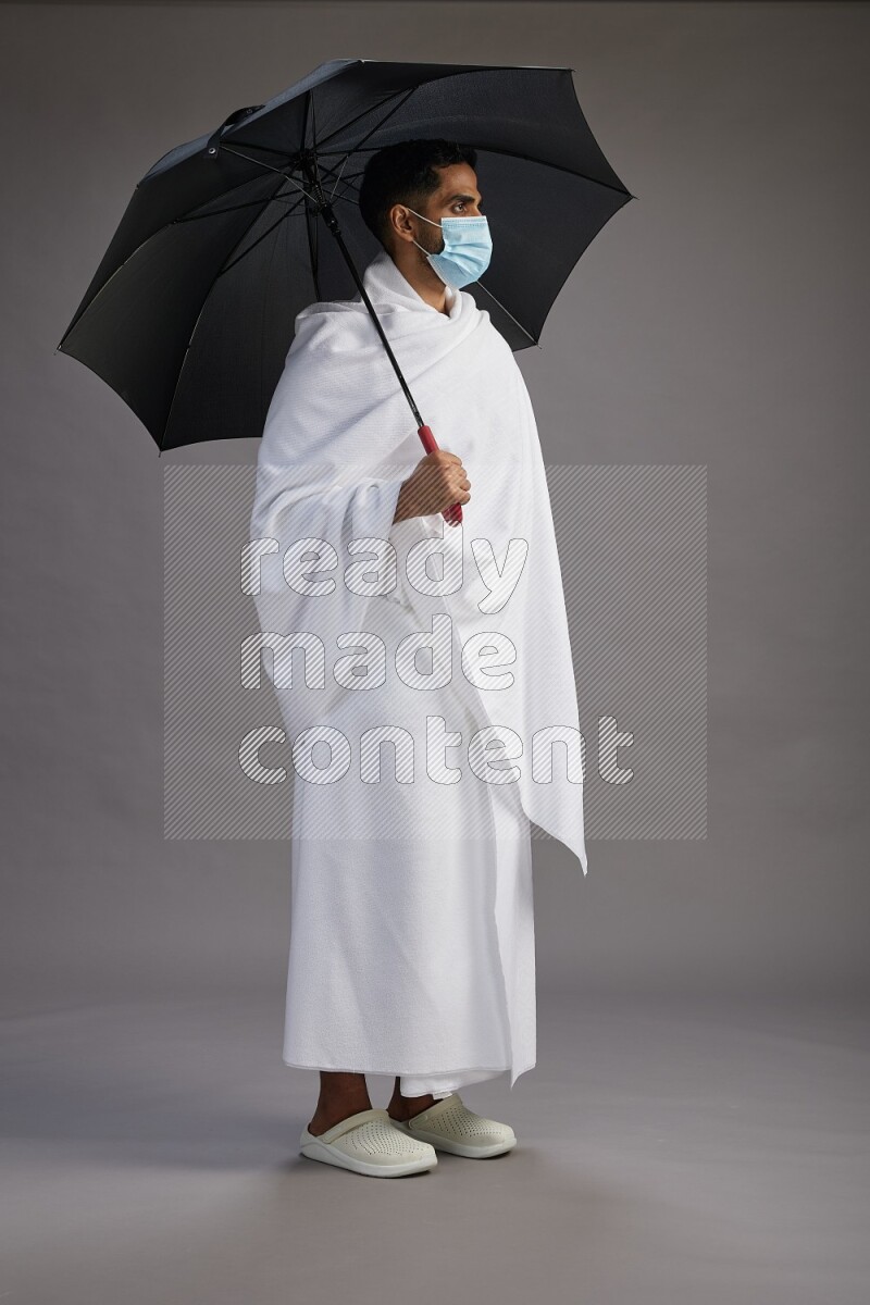 A man wearing Ehram and mask Standing holding umbrella on gray background on gray background
