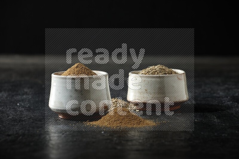 2 beige bowls, one full of cumin powder and the other full of seeds on a textured black flooring