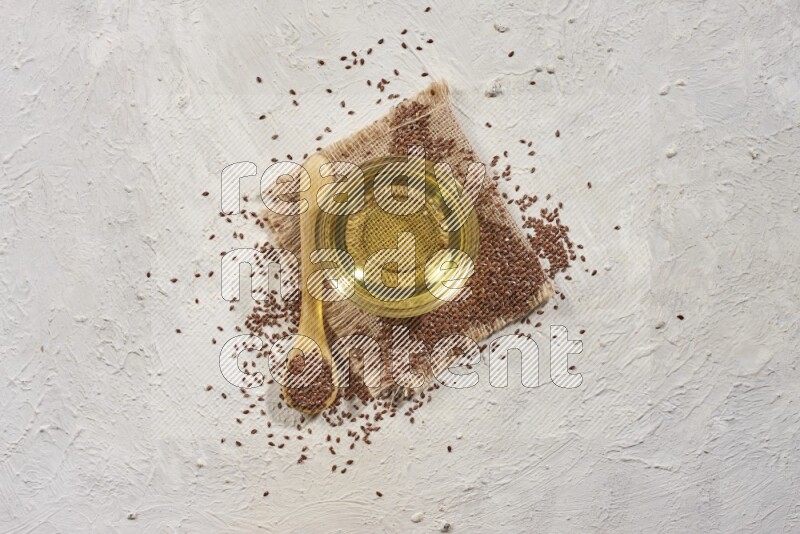A glass bowl full of flaxseeds oil and wooden spoon full of flaxseeds with spread seeds on burlap fabric on a textured white flooring