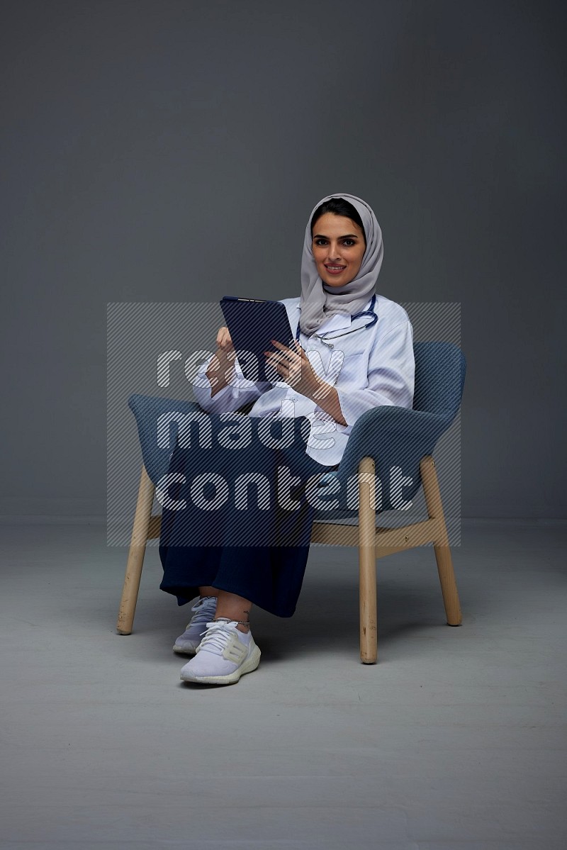 A female doctor wearing a light gray head scarf sitting on a dark grey chair holding an electronic device and pointing to different directions eye level on a grey background
