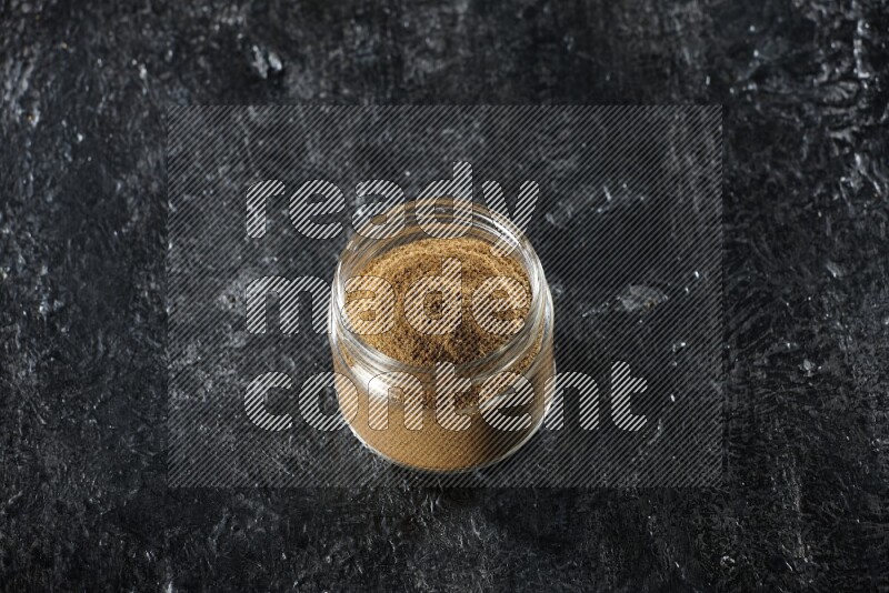 A glass jar full of cumin powder on a textured black flooring