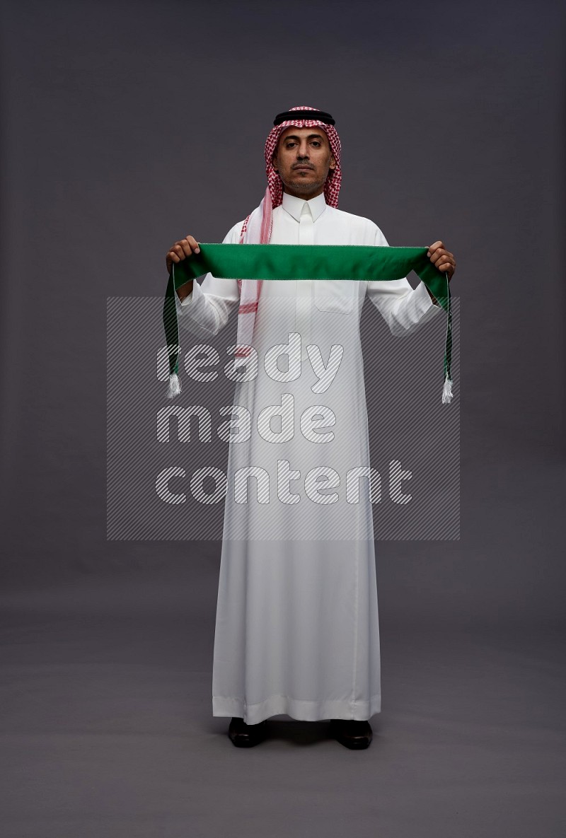 Saudi man wearing thob and shomag standing holding Saudi flag on gray background