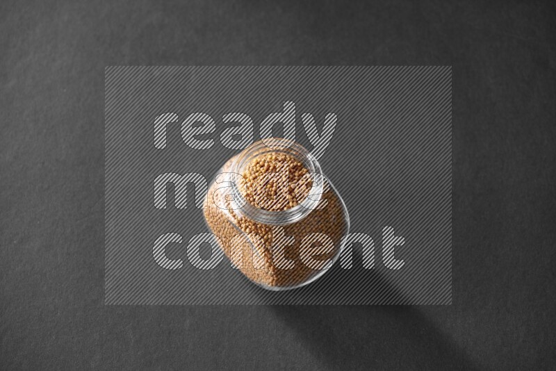 A glass spice jar full of mustard seeds on black flooring