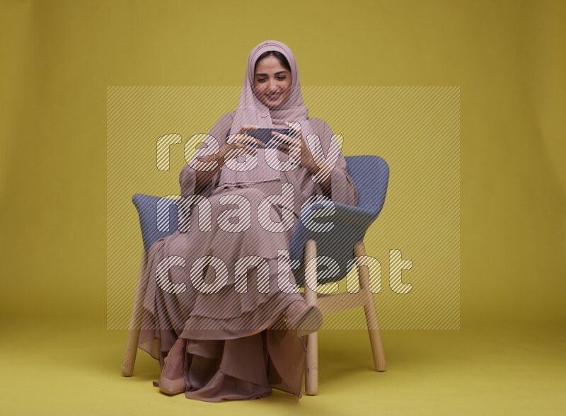 A woman Playing Game Sitting  on a Yellow Background wearing Brown Abaya with Hijab