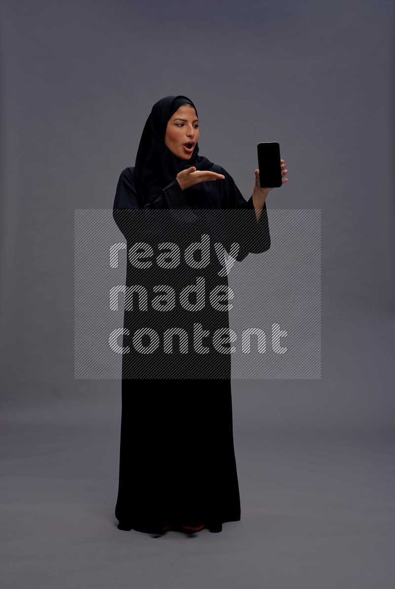 Saudi woman wearing Abaya standing showing phone to camera on gray background