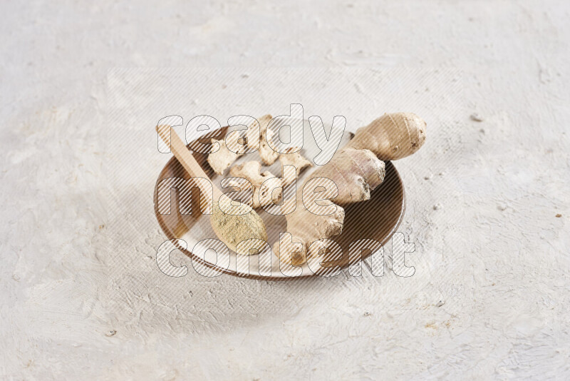 A wooden spoon full of ground ginger powder with fresh and dried ginger, all on a pottery plate on white background