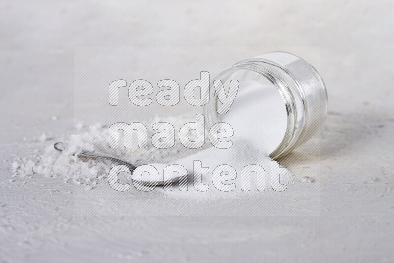 A glass jar full of table salt with some sea salt crystals beside it on a white background