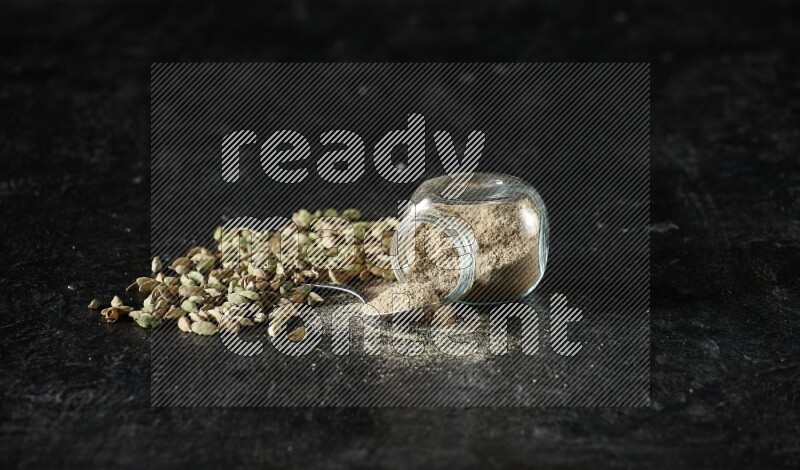A flipped glass spice jar and a metal spoon full of cardamom powder and cardamom seeds spreaded on textured black flooring