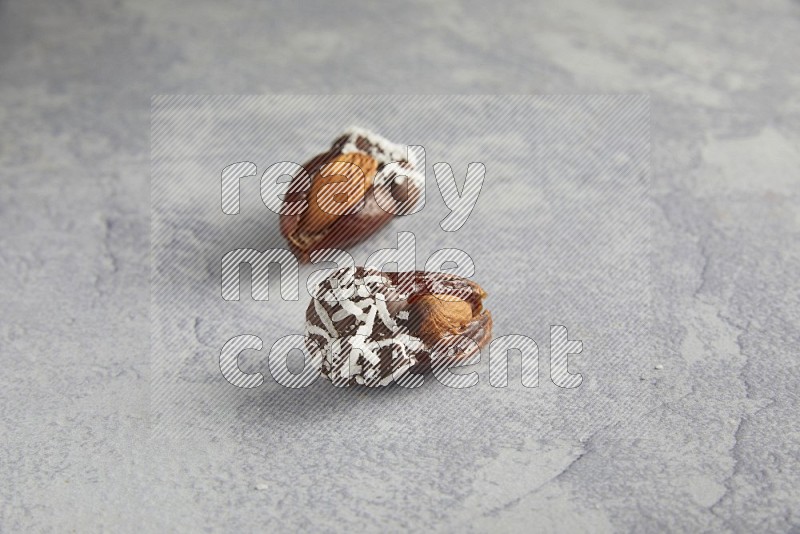 Two Almond stuffed dates covered with dark chocolate and coconut on a light grey background