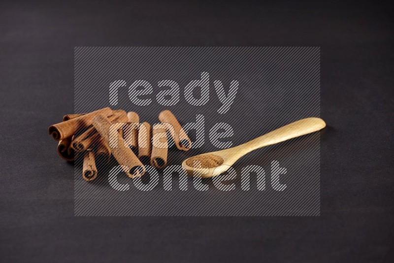Cinnamon sticks stacked beside a wooden spoon full of cinnamon powder on black background