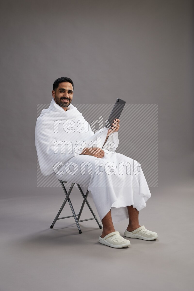 A man wearing Ehram sitting on chair holding a tablet on gray background