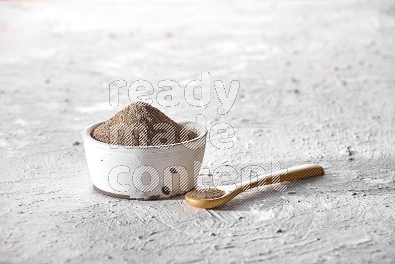 A pottery white bowl full of black pepper powder and wooden spoon full of powder on textured white flooring