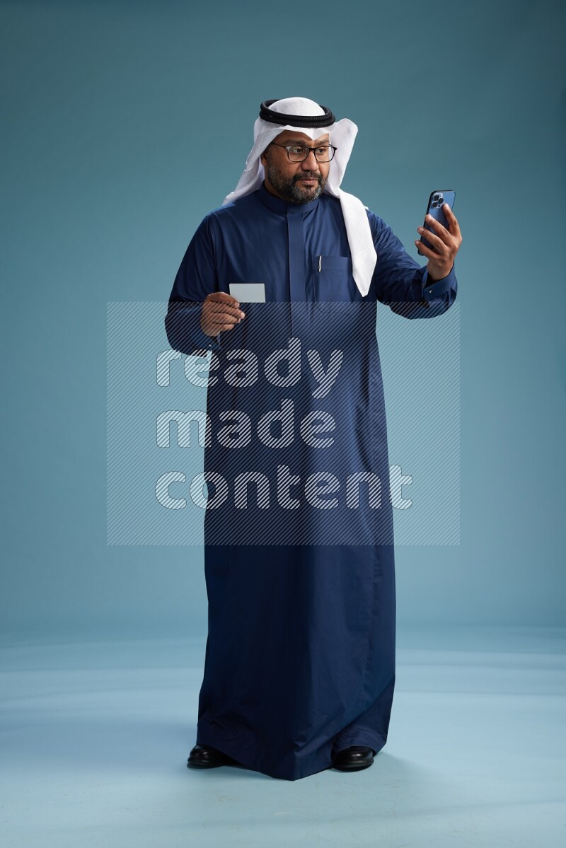 Saudi Man with shimag Standing holding ATM card while talking on phone on blue background