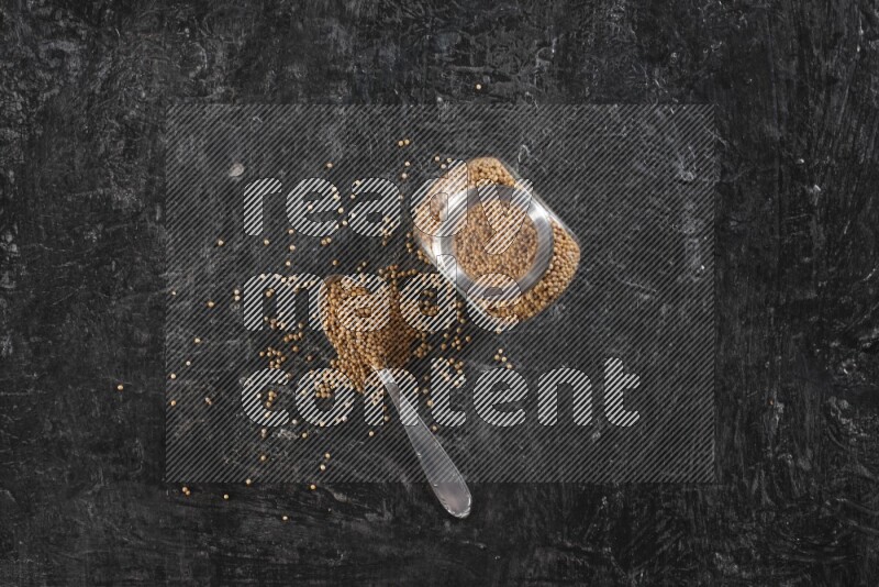 وعاء زجاجي وملعقة معدنية كلاهما ممتلئ ببذور الخردل على أرضية سوداء