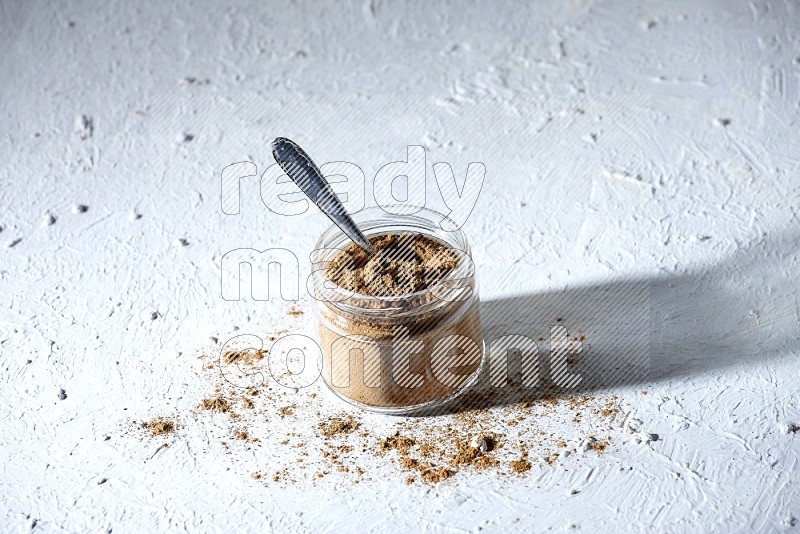 A glass jar and a metal spoon full of allspice powder on a textured white flooring