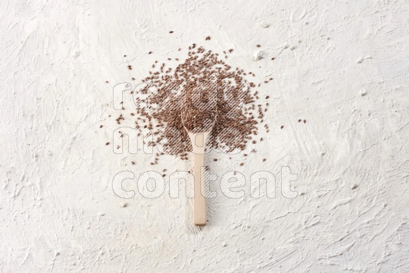 A wooden spoon full of flax seeds surrounded by flax seeds on a textured white flooring