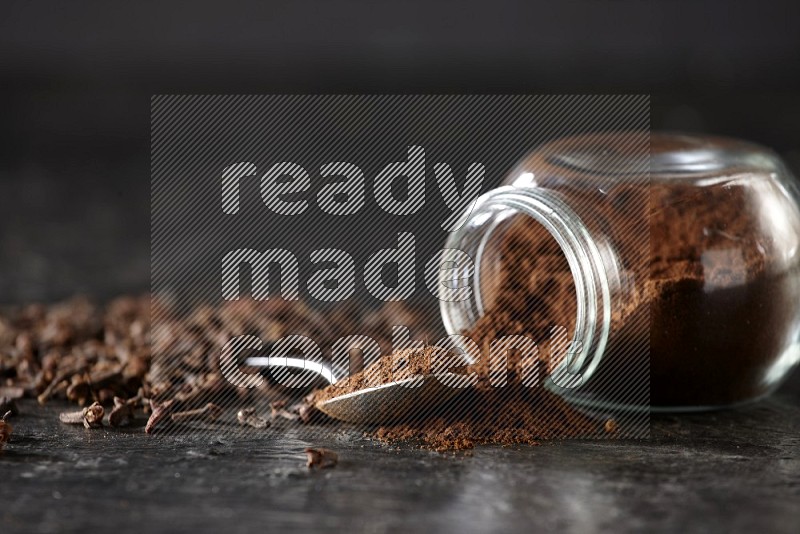 A flipped glass spice jar and a metal spoon full of cloves powder with cloves spread on textured black flooring