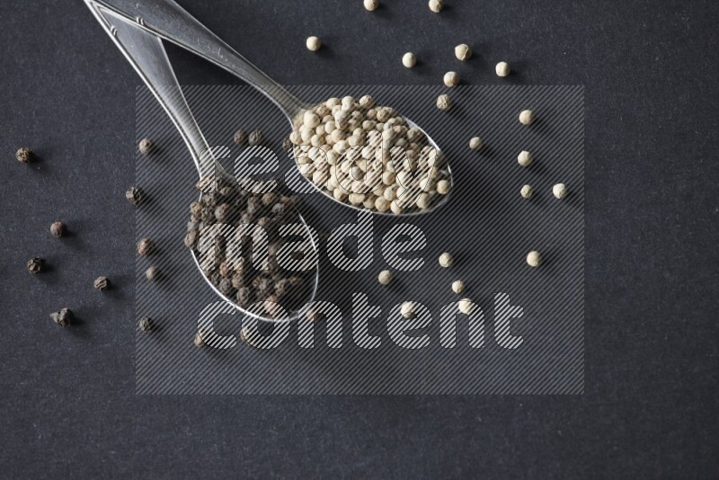 ملعقتان معدنيتان ممتلئتان بحبوب الفلفل الأسود والأبيض مع حبوب متناثرة على أرضية سوداء