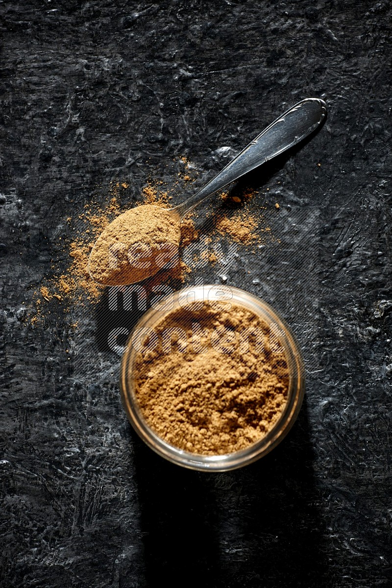 A glass jar and a metal spoon full of allspice powder on a textured black flooring