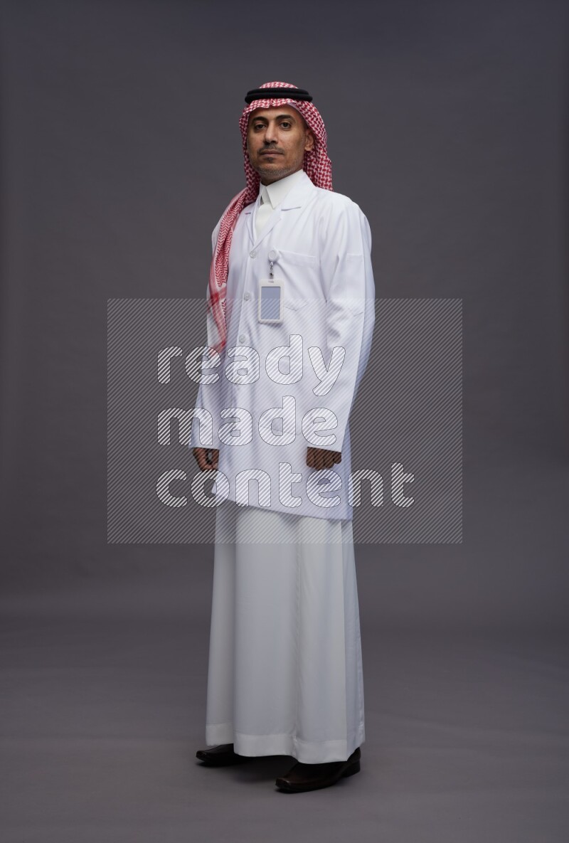 Saudi man wearing thob with lab coat and shomag with pocket employee badge standing interacting with the camera on gray background