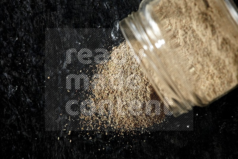A flipped glass jar full of cardamom powder and powder spilled out of it on textured black flooring