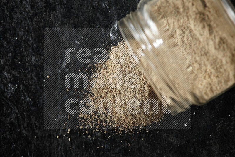 A flipped glass jar full of cardamom powder and powder spilled out of it on textured black flooring