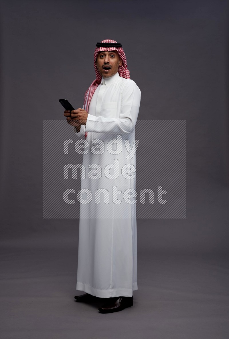 Saudi man wearing thob and shomag standing texting on phone on gray background